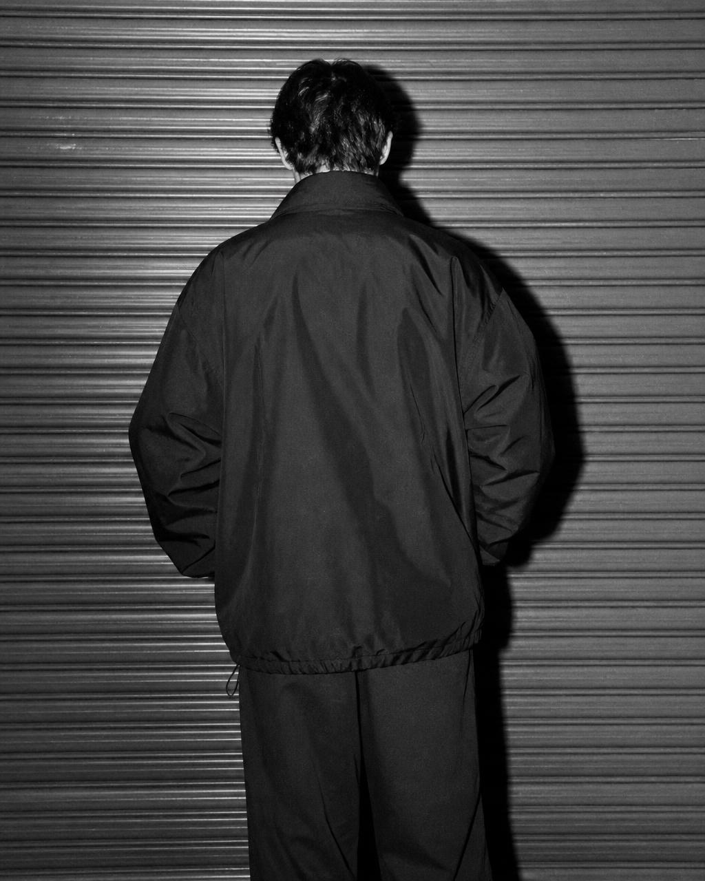 Tampak belakang model mengenakan coach jacket hitam di depan rolling door
